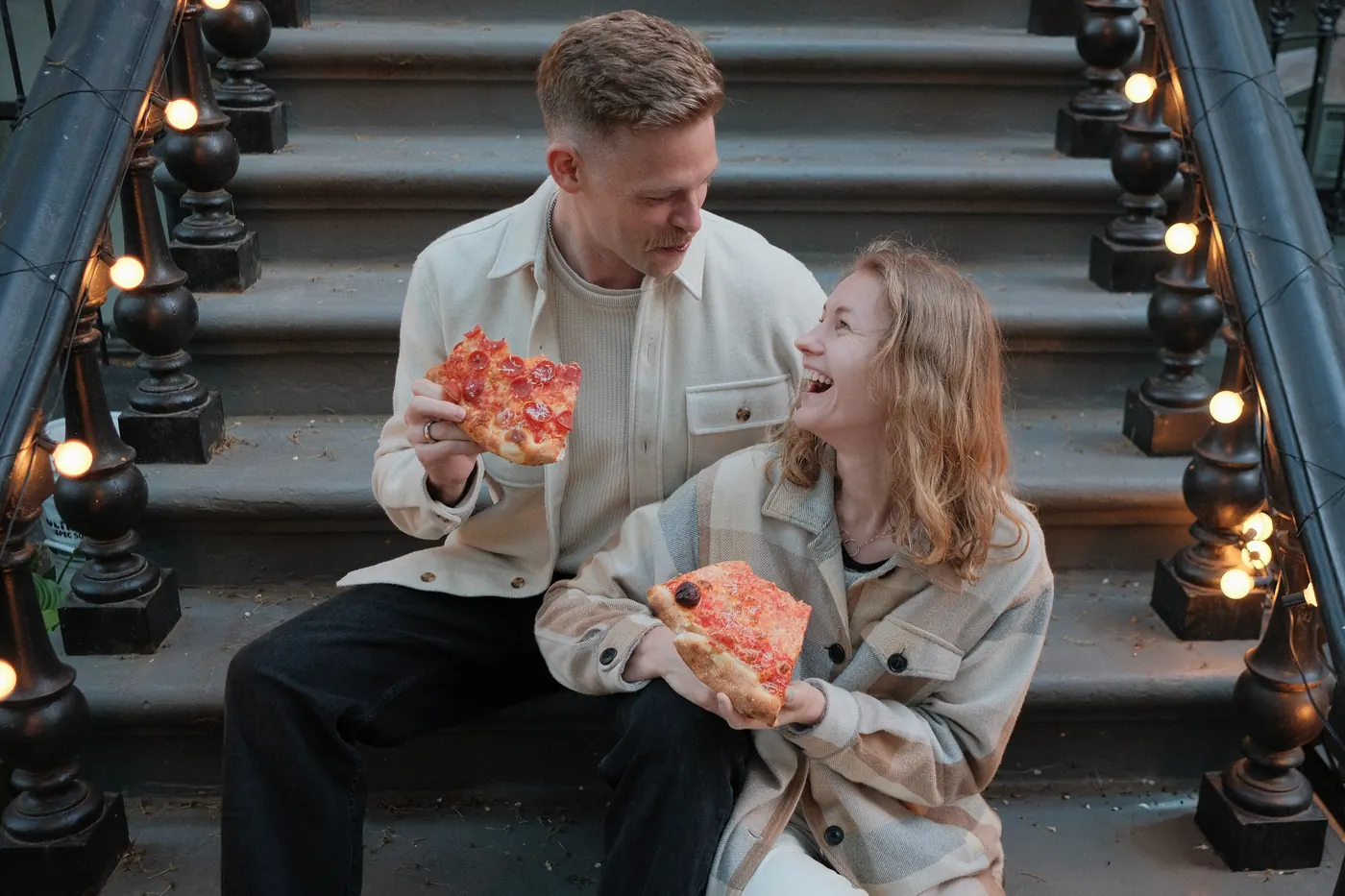 Couple laughing over pizza slices on stoop steps with string lights
