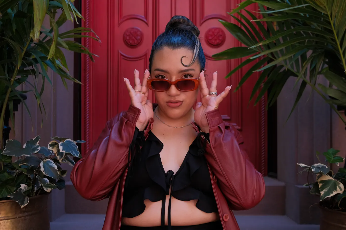 Woman in a red leather jacket adjusting orange sunglasses in front of a red door